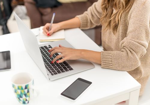 Een vrouw die aan een bureau zit typend op haar laptop en schrijft in haar schrift.